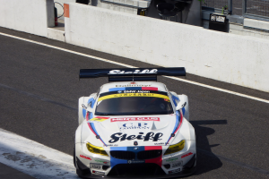 Ein BMW M6 GT3 Rennauto auf einer Rennstrecke mit einer Wand, einem Maschendrahtzaun und anderen Gegenständen im Hintergrund.