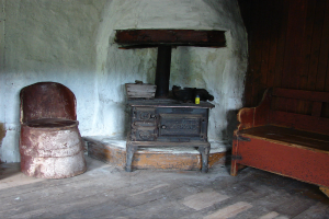 Ein altmodischer Kachelofen in einem Raum mit einer hölzernen Bank auf der rechten Seite, einem Stuhl auf der linken Seite und einer Wand im Hintergrund.