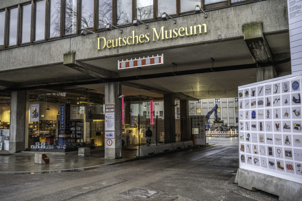 Außenansicht des Deutschen Museums in Berlin, das eine große Glasfassade mit Säulen, ein Namensschild, ein Seiteninformationsschild mit Bildern und Text sowie umliegende Gebäude, Bäume und Himmel zeigt.