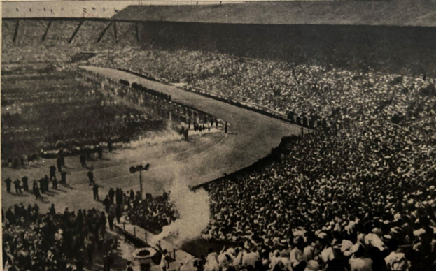 Schwarz-weißes Foto einer großen Menschenmenge in einem Stadion mit einer zentralen Straße und Pfosten auf beiden Seiten, mit Text unten.