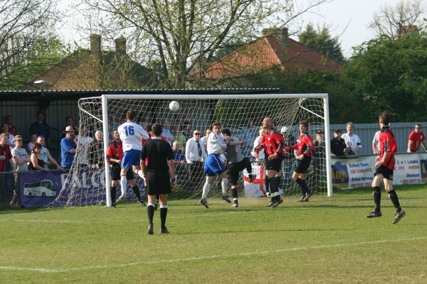 Spieler spielen Fußball auf einem Feld mit einem Tor hinter ihnen, während Zuschauer stehen und zuschauen, mit Bäumen und Häusern im Hintergrund.