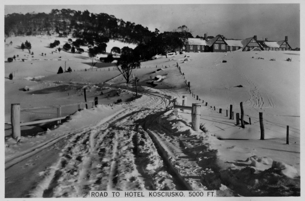 Ein Schwarz-Weiß-Foto einer verschneiten Straße, die zu einem Hotel führt, mit Pfälen, Bäumen und Häusern im Hintergrund und Text unten.