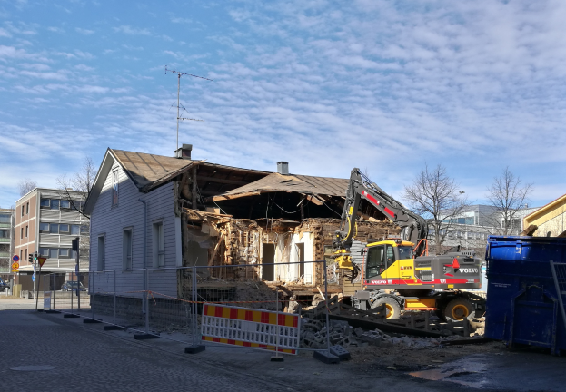 Eine Baustelle mit einem großen Gebäude, das abgerissen wird, mit einem Kran, einem Zaun, einer Absperrung, Steinen, einem Container, Bäumen, Pfosten, einem Schild, einem Straßenpfosten, Fahrzeugen, Gebäuden mit Fenstern und einem bewölkten Himmel.