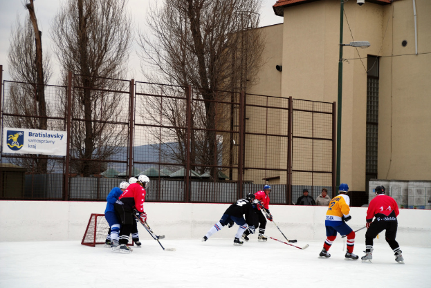 Menschen beim Eishockeyspielen auf einer Eisfläche mit Gebäuden, Bäumen, einer Straßenlaterne, einem Namensschild und Zäunen im Hintergrund unter dem Himmel.