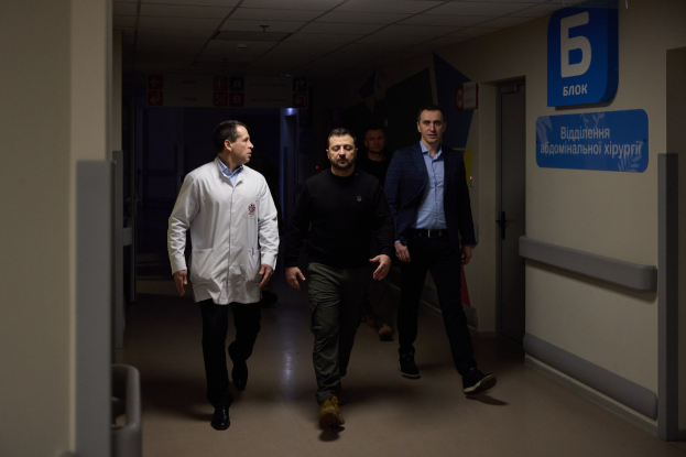 Eine Gruppe von Männern geht einen hell erleuchteten Krankenhausflur entlang, mit einer Tür auf der rechten Seite und Texttafeln auf der linken Seite, die besorgt aussehen.