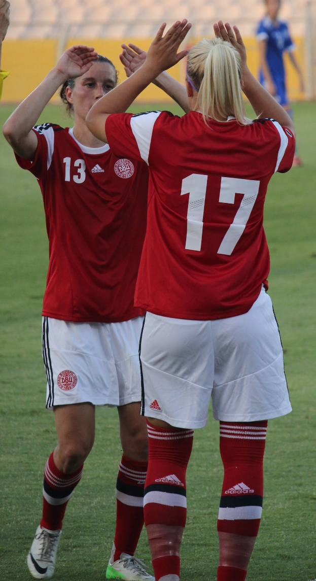 Zwei Frauen in roten T-Shirts und weißen Shorts klatschen auf einem Fußballfeld mit einer Person in einem blauen Kleid und einer gelben Tafel im Hintergrund.