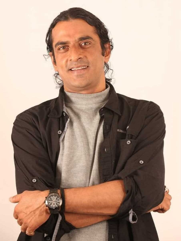 Ein Mann in einem schwarzen und aschfarbenen Kleid, der eine Uhr tr├Ągt, steht mit überschlagenen Armen vor einem wei├čen Hintergrund und sieht nachdenklich aus.