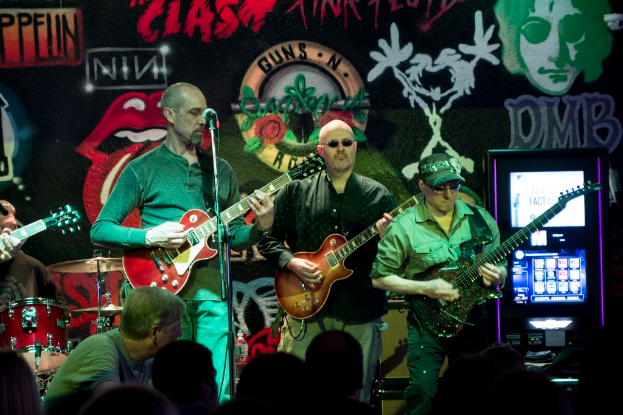 Drei Männer auf einer Bühne mit Gitarren, umgeben von einer Menge, mit einer bemalten Wand im Hintergrund.