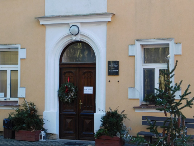 Eine hölzerne Tür mit einem Kranz, flankiert von Fenstern, steht vor einem Gebäude mit einer Bank in der Nähe, umgeben von Zimmerpflanzen, einer Lampe und anderen verstreuten Gegenständen.