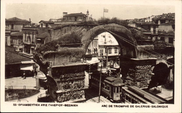 Schwarz-weiß-Fotografie einer Stadtstraße mit einer Bogenbrücke in der Mitte, umgeben von Gebäuden, Bäumen, Pfählen, Drähten und Menschen, mit Text unten.