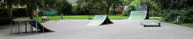 Ein Park mit einer Skateboard-Rampe, umgeben von Bänken, Geländern, Gras, Pflanzen, Bäumen und Gebäuden im Hintergrund.
