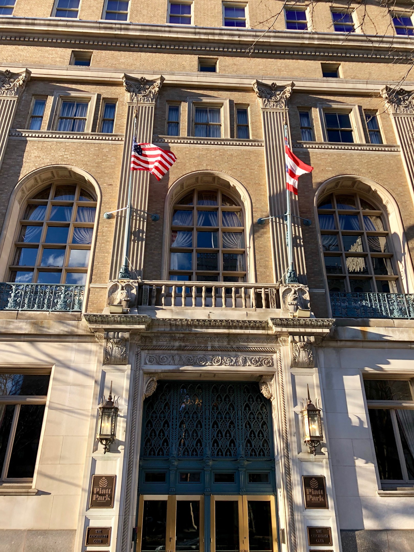 Außenansicht des Plaza Hotels in New York City, mit Glasfenstern, Türen, Säulen, Geländern, Leuchten, Texttafeln und Flaggen vor einem Himmelhintergrund.