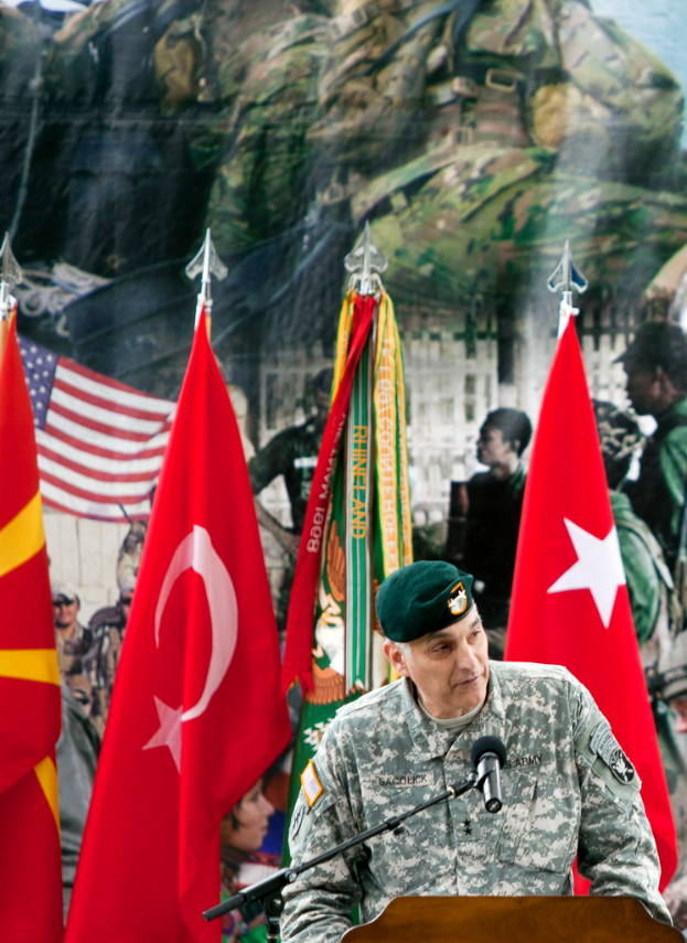 Ein Soldat steht an einem Podium mit einem Mikrofon, mit verschiedenen Flaggen und einem Plakat mit Militärpersonal im Hintergrund.