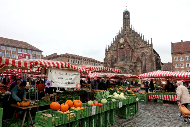 Ein belebter Markt in Nürnberg, Deutschland, mit einer Vielfalt an Obst und Gemüse, Menschen mit Taschen, Zelten, Gebäuden mit Fenstern, einem Uhrenturm und sichtbarem Himmel.