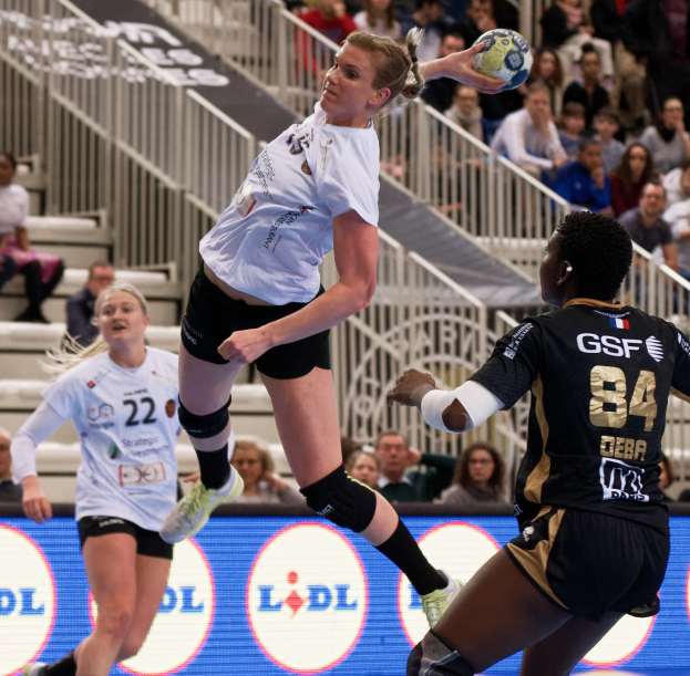 Eine Gruppe von Frauen, die Handball auf einem Court spielen, mit einem Ball in der Luft, Zuschauern im Hintergrund und einem "Handball-Weltmeisterschaft 2019 - Tag 2"-Schild.