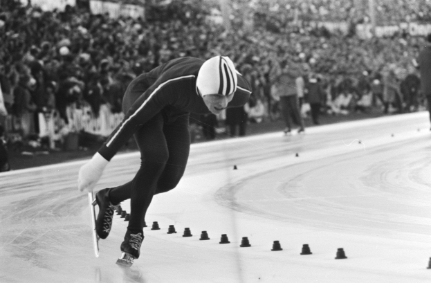 Ein Schwarz-Weiß-Foto eines Eisschnellläufers in Aktion auf dem Eis, der einen Helm und Eislaufschuhe trägt und im Hintergrund einige unscharfe Personen zu sehen sind.