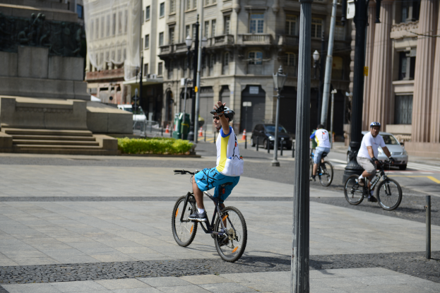 Drei Männer fahren mit Fahrrädern auf einer Straße mit Gebäuden und Straßenlaternen im Hintergrund und zwei Autos auf einer Straße rechts.