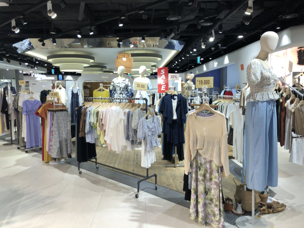 Ein Schaufenster einer Kleiderboutique mit Schaufensterpuppen in Kleidern an Bügeln, beleuchtet von Deckenlampen und umgeben von Hängeschildern und Bodenartikeln.