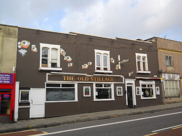 Eine Straße und ein Fußweg unten, mit Gebäuden in der Mitte mit Türen, Fenstern und dem Text "Der alte Stapel"; eine rote Wand und eine Glastür links, ein lila Schild mit Text und den Himmel oben.