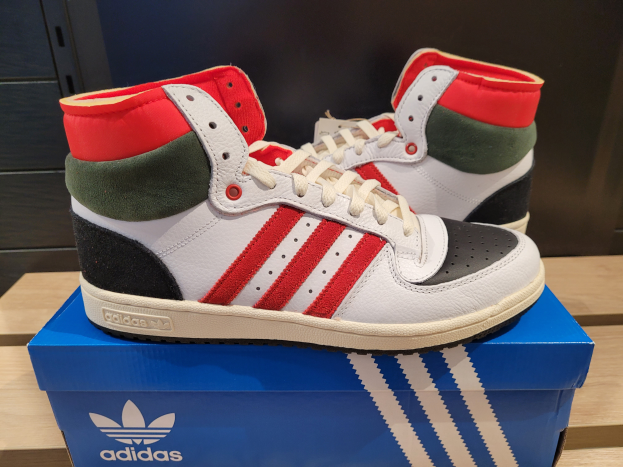 Ein Paar weiße, rote, grüne und schwarze Adidas Originals Top Ten Hi Schuhe auf einer blauen und braunen Kartonbox, die auf einem cremefarbenen Tisch steht, mit einer schwarzen Wand im Hintergrund.