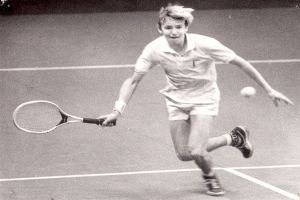 Ein junger Mann in weißer Tennisbekleidung schlägt einen Ball auf einem Tennisplatz mit einem entschlossenen Gesichtsausdruck.