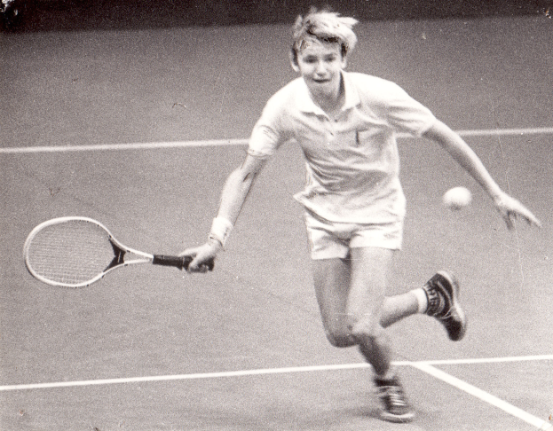 Ein junger Mann in weißer Tennisbekleidung schlägt einen Ball auf einem Tennisplatz mit einem entschlossenen Gesichtsausdruck.