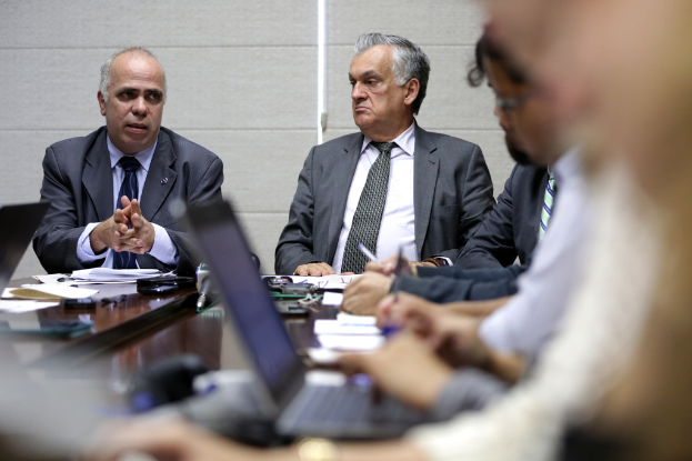 Ein Mann in einem blauen Hemd und Blazer steht in der Ecke eines Konferenzraums und spricht mit vier sitzenden Männern, mit einem Tisch voller Gegenstände vor ihnen und einer weißen Wand dahinter.