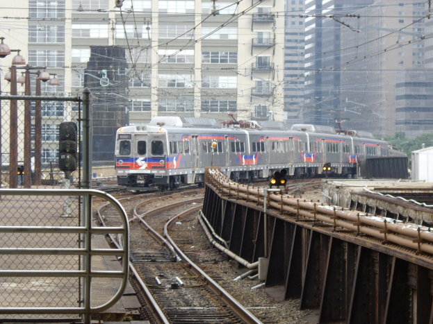 Ein Nahverkehrszug fährt auf Schienen neben hohen Gebäuden, mit Eisenbahninfrastruktur, Bäumen und anderen Objekten im Hintergrund.