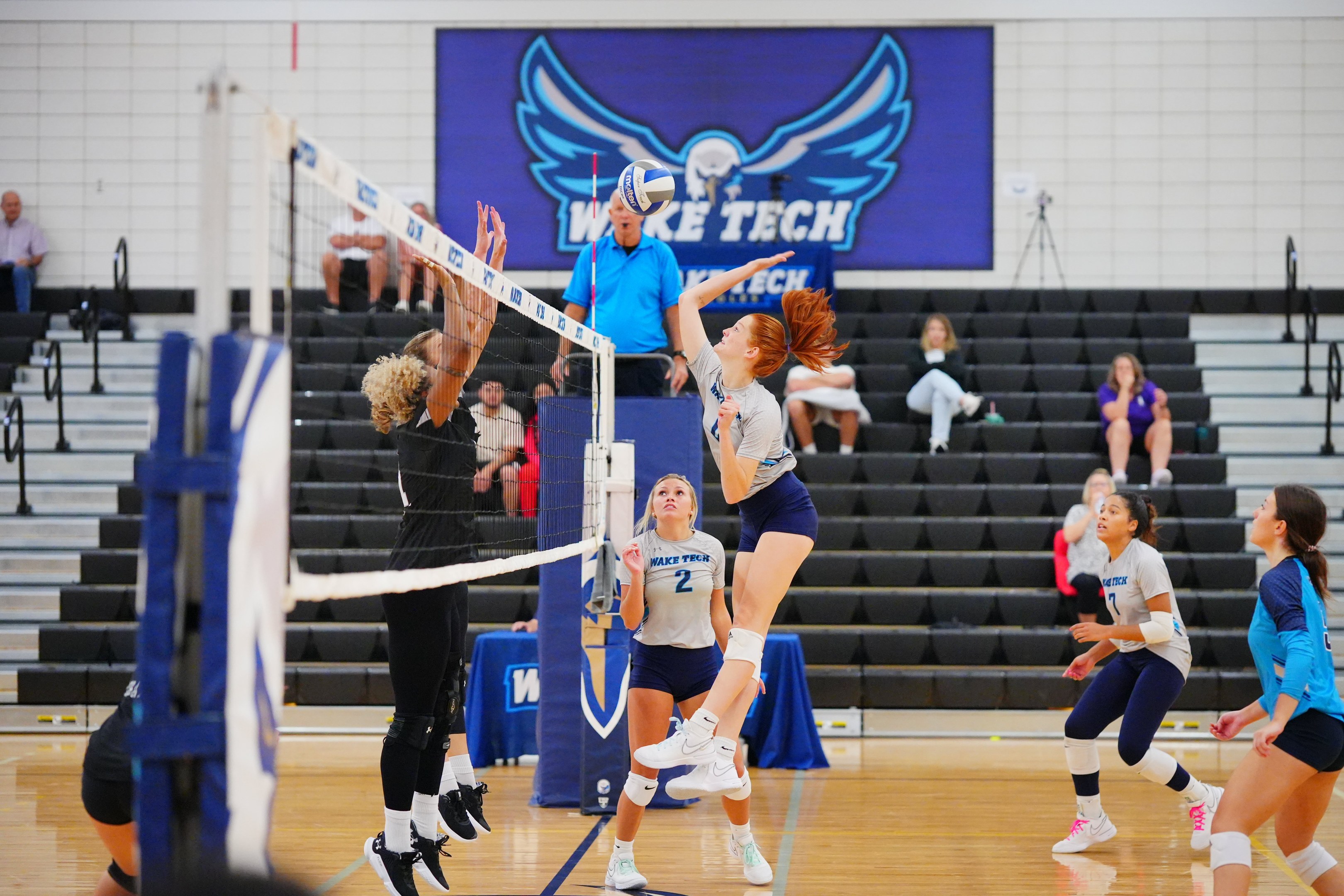 Frauen beim Volleyballspielen mit einem Netz, Zuschauern auf einer Treppe und einem Meisterschaftsbanner mit einem Adler an der Wand.