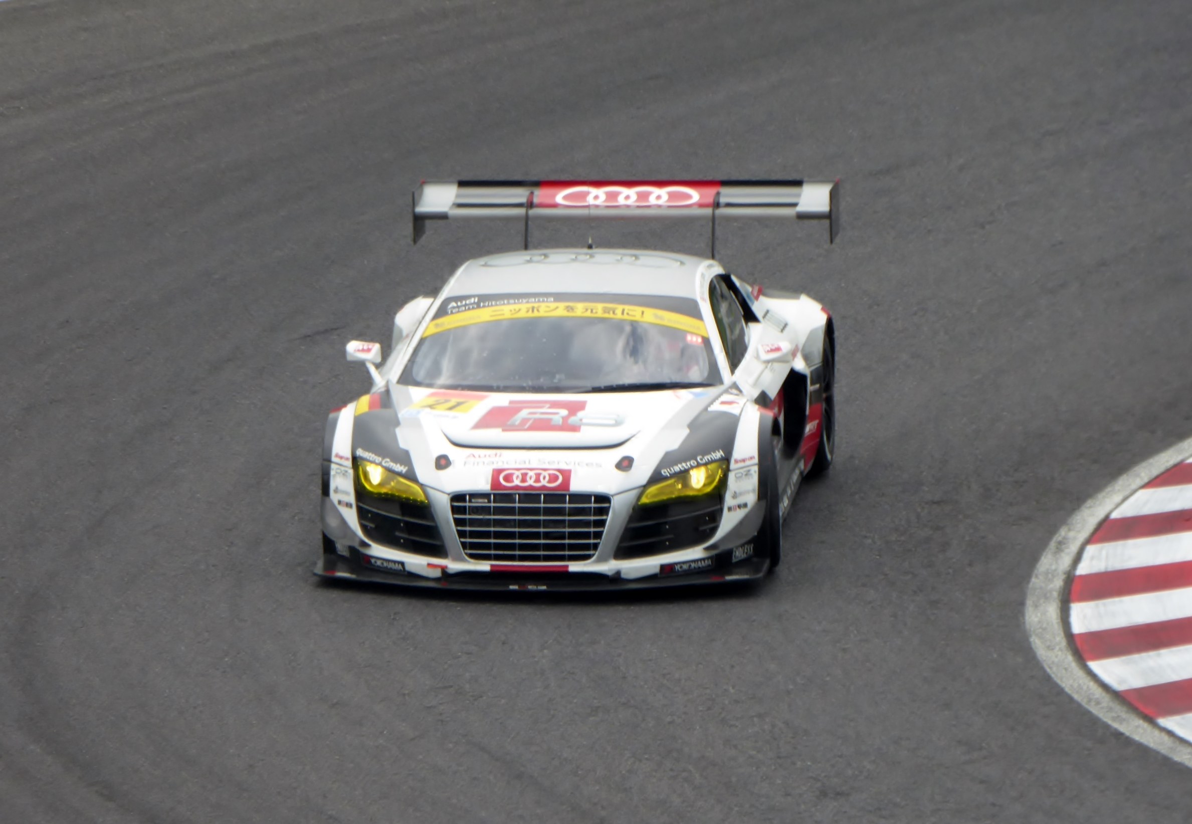 Ein leuchtend roter und weißer Audi R8 LMS GT3 Rennauto mit dem Audi-Logo auf der Seite, gefahren von einer Person in einem schwarzen Rennanzug und Helm, der um eine Rennstrecke fährt.
