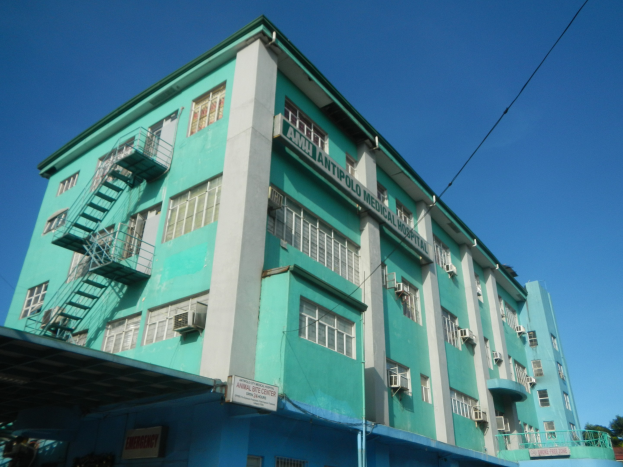 Großes grünes Gebäude mit zahlreichen Fenstern, Klimaanlagen, Treppen, Geländern, Schilden, Strommasten, Stromkabeln, Fahrzeugen auf der Straße, Bäumen und einem klaren blauen Himmel.