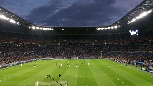 Ein Fußballspiel wird in einem großen Stadion mit Menschen auf dem Rasen, einem Tor, einer Kamera mit Stativ, Bannern mit Text, einem Dach mit Deckenleuchten und einem Anzeigebildschirm unter einem bewölkten Himmel gespielt.
