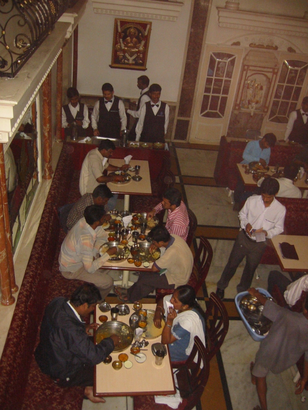 Eine Gruppe von Menschen an Tischen sitzend, die essen, mit einigen im Hintergrund stehend, die servieren, ein Foto mit Blumen auf der Wand und eine Tür im Hintergrund.