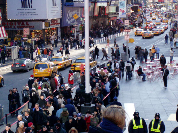 Fahrzeuge fahren auf einer Straße mit Fußgängern auf beiden Seiten, neben Stühlen, Tischen, Laternen, Schildern und Ampeln sowie Gebäuden und Geschäften auf beiden Seiten.