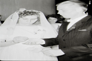 Ein Schwarz-Weiß-Foto von einer Braut und einem Bräutigam, die sich die Eheringe reichen, mit der Braut in einem weißen Kleid und Schleier und dem Bräutigam in einem Anzug, mit ein paar Leuten im Hintergrund.