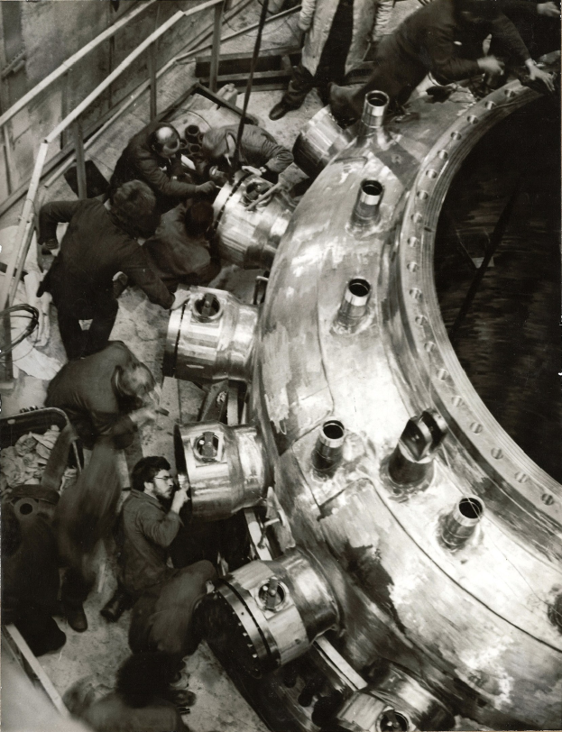 Schwarzes und weißes Bild von Männern, die an einem großen Metallreaktor in einer industriellen Fabrikumgebung arbeiten, mit Treppen, Geländern und einer Wand im Hintergrund.