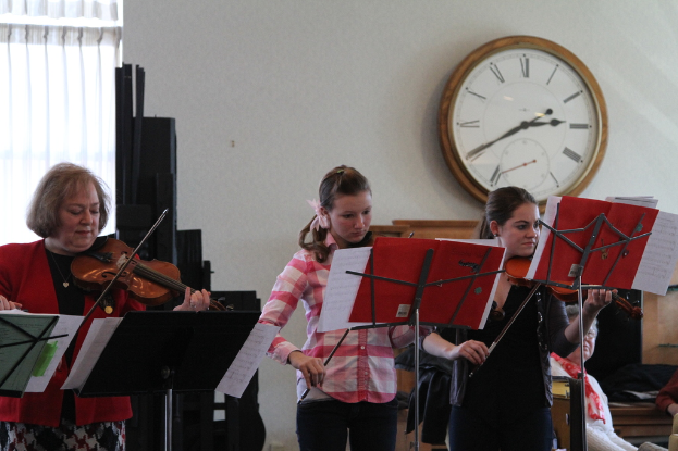 Drei Frauen, die Geigen auf der Bühne spielen, mit Notenpulten und Notenblättern davor, während ein Zuschauer im Vordergrund sitzt; eine Uhr hängt an der weißen Wand im Hintergrund, und ein weißer Vorhang ist links sichtbar.