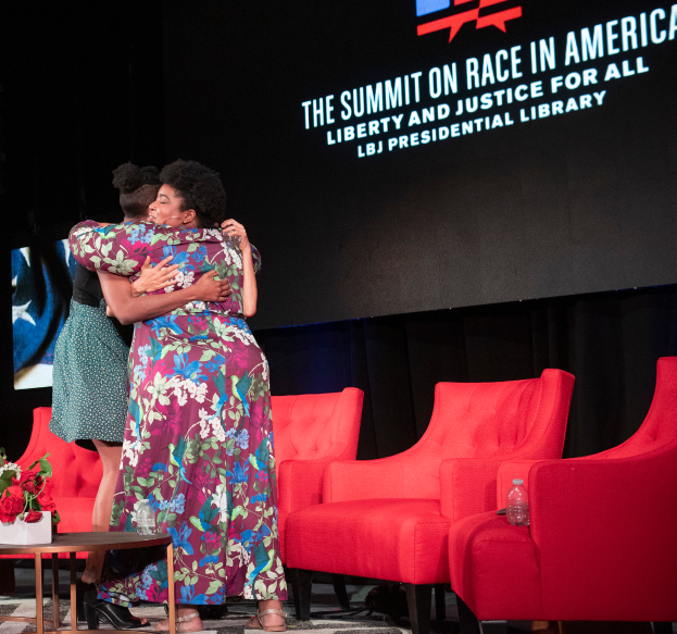 Zwei Frauen, identifiziert als L.B.J. Präsidentin und First Lady Michelle Obama, umarmen sich auf einer Bühne mit einem Bildschirm, der 'The Summit on Race in America' anzeigt. Ein Tisch mit einer Vase und einer Flasche ist links und rote Stühle rechts.