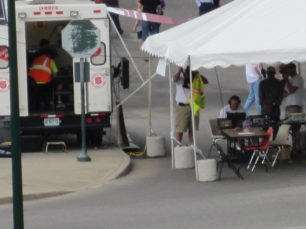 Ein Rettungswagen steht links neben einer Straße, während rechts ein Zelt viele Menschen schützt, bei denen ein Tisch und ein Stuhl in der Nähe stehen.