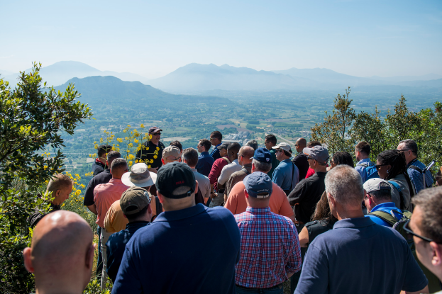 Eine Gruppe von Menschen auf einem Berg, umgeben von Bäumen und Hügeln, mit einem klaren blauen Himmel im Hintergrund, einige tragen Mützen.