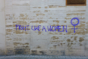 Eine Wand mit den Worten "Kämpfe wie eine Frau" in Blau gesprüht, mit einem Fenster oben und einem Boden unten.