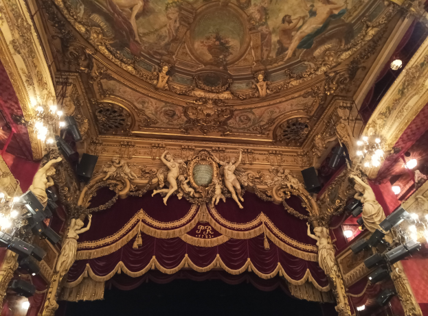 Theaterinnenraum mit roter Gardine, Kronleuchtern, Skulpturen, Deckenmalerei und einer Bühne mit einigen Menschen unten.