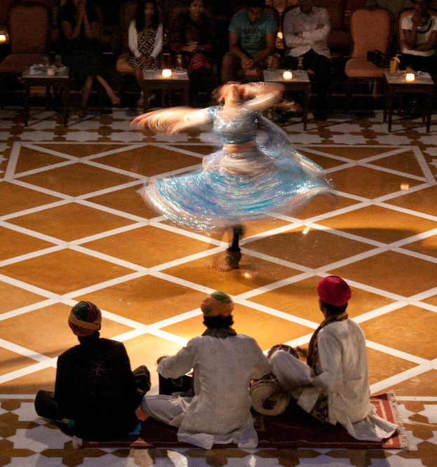 Eine Gruppe von Menschen sitzt im Hintergrund auf Stühlen und spielt Musikinstrumente, während eine Person in der Mitte tanzt und von Hockern mit Kerzen umgeben ist.