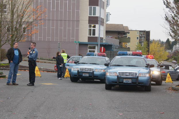 Autos auf einer Stra√üe mit vier Personen in der N√§he, Geb√§ude mit Fenstern im Hintergrund, B√§ume und Warnkegel.