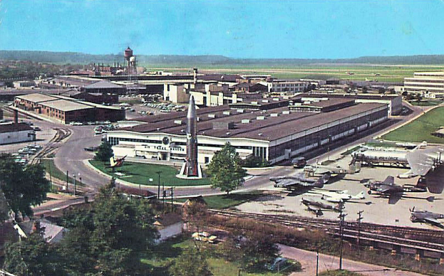 Ein altes Foto eines Flughafens mit geparkten Flugzeugen, Gebäuden, Bäumen, Straßen, Fahrzeugen, Masten, Gras, Bahnschienen, Türmen und Hügeln unter einem klaren blauen Himmel, mit Text unten.