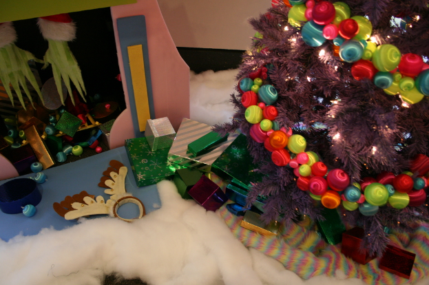 Eine Indoor-Szene mit einem geschmückten Weihnachtsbaum auf der rechten Seite, Geschenkboxen und Dekorationsgegenständen in der Mitte und einer Hütte mit Dekorationen oben links, mit Watte und Wolle am Boden.