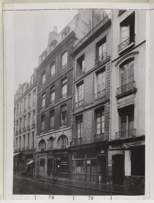 Schwarzes und weißes Foto einer Stadtstraße mit Gebäuden, die Fenster, Geländer und Namensschilder sowie eine Straße unten zeigen.