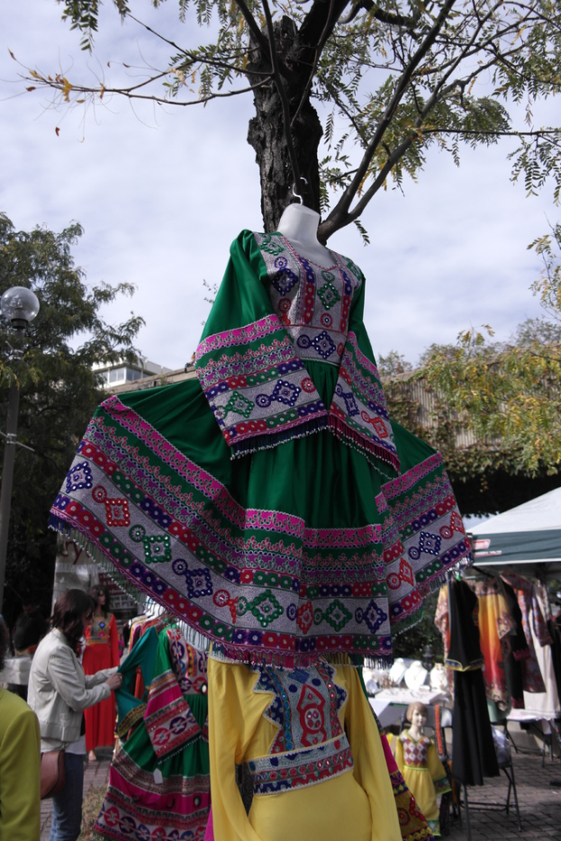 Eine Schaufensterpuppe steht inmitten eines belebten Einkaufsgebiets mit mehreren Geschäften, einer Person, die Kleider an einem Ständer arrangiert, einem Gebäude und Bäumen im Hintergrund.