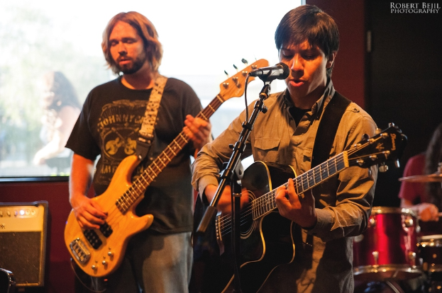 Eine Person auf einem Boden stehend, die eine Gitarre hält und singt, neben sich eine weitere Person, die Gitarre spielt und im Hintergrund ein Schlagzeug.