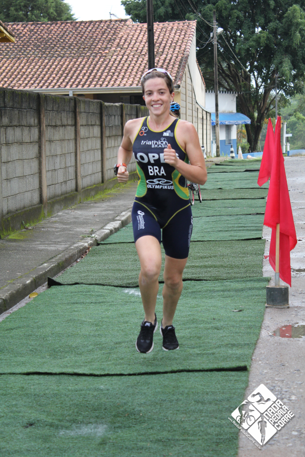 Eine Frau in einem Triathlon-Anzug läuft strahlend eine Straße entlang, mit einer roten Fahne auf einem Mast rechts und Häusern, Bäumen, Pfählen, Drähten und einem klaren blauen Himmel im Hintergrund.
